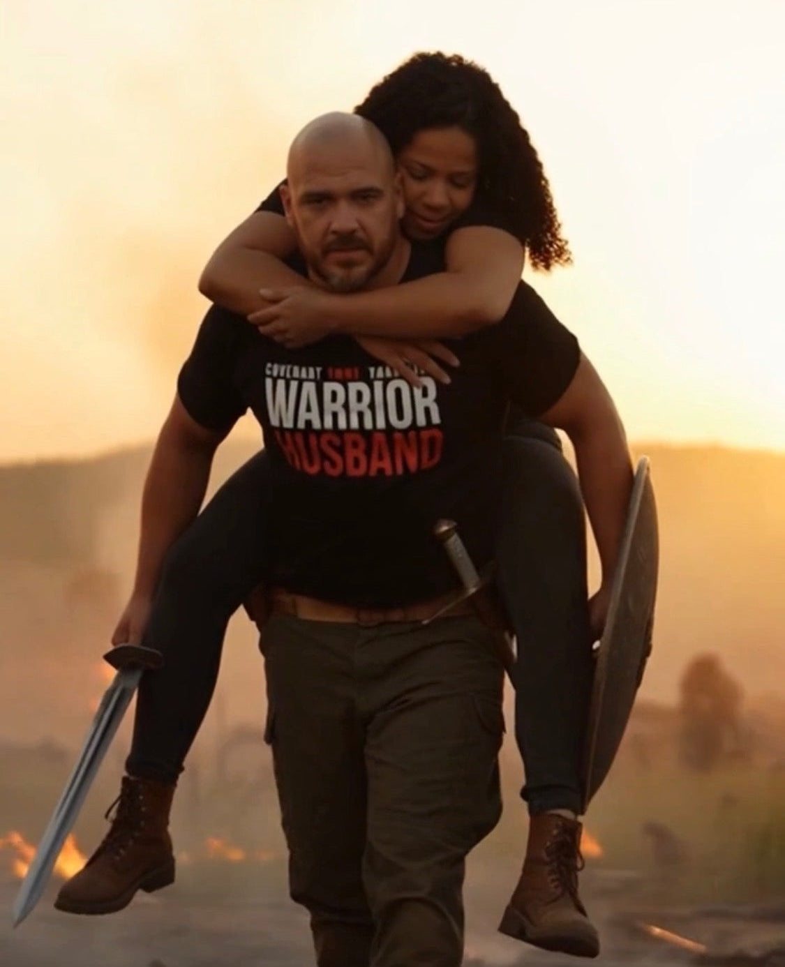 Man carrying a woman on his back with 'Warrior Husband' shirt in a sunset setting